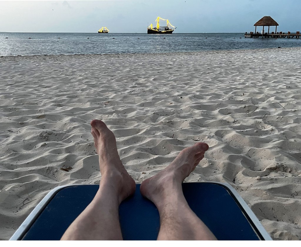Person lying on a beach - feet shown at the end of a chaise lounge with the ocean in the distance